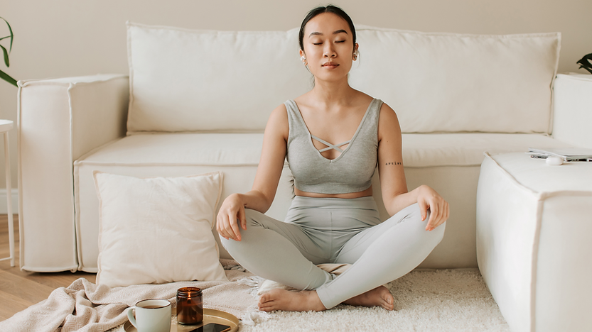 Uma mulher sentada no chão em posição de meditação, com as pernas cruzadas e as mãos repousando sobre os joelhos. Ela está em um ambiente claro e tranquilo, refletindo um momento de paz e introspecção.