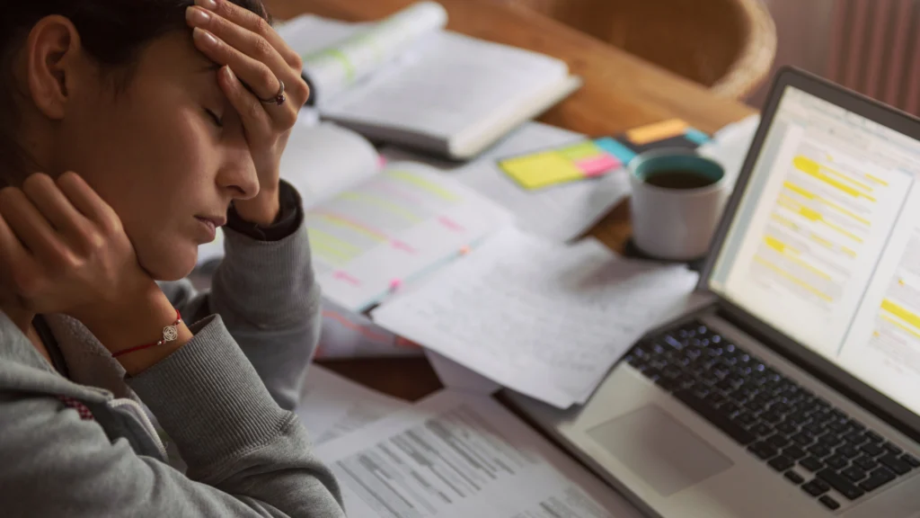 Pessoa sentada à mesa com a mão na testa, aparentando cansaço e estresse, cercada de papéis, livros e um laptop aberto. Representa sinais de burnout e ansiedade, mostrando sobrecarga de trabalho e dificuldade de concentração.